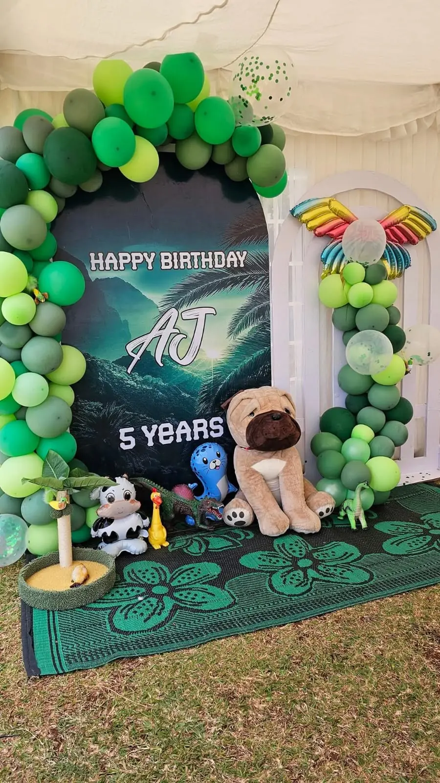 A green animal-themed fifth birthday backdrop with balloons and plush animal decorations