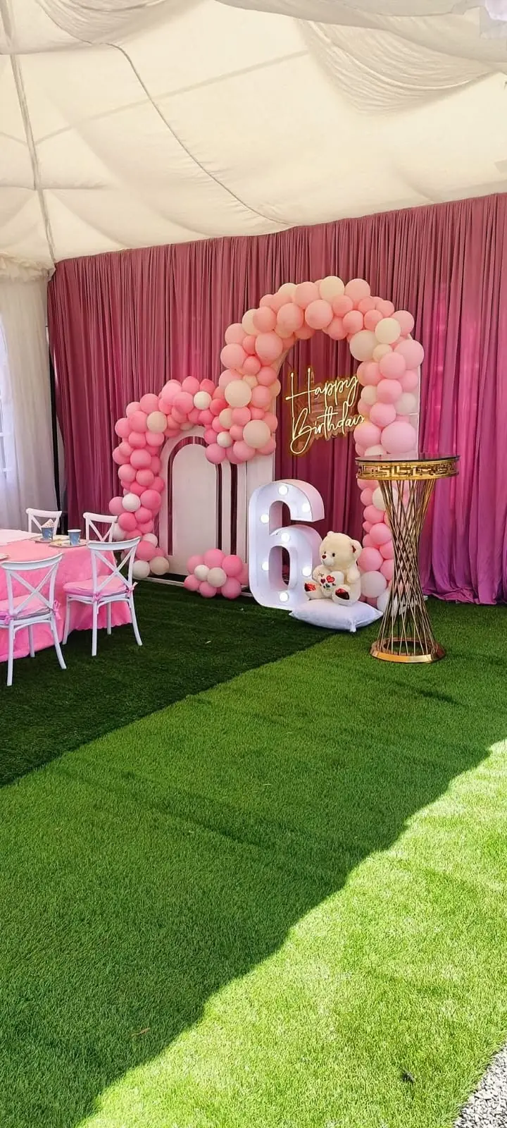 A pink sixth birthday setup with balloon arches, an illuminated number and a dressed children's table