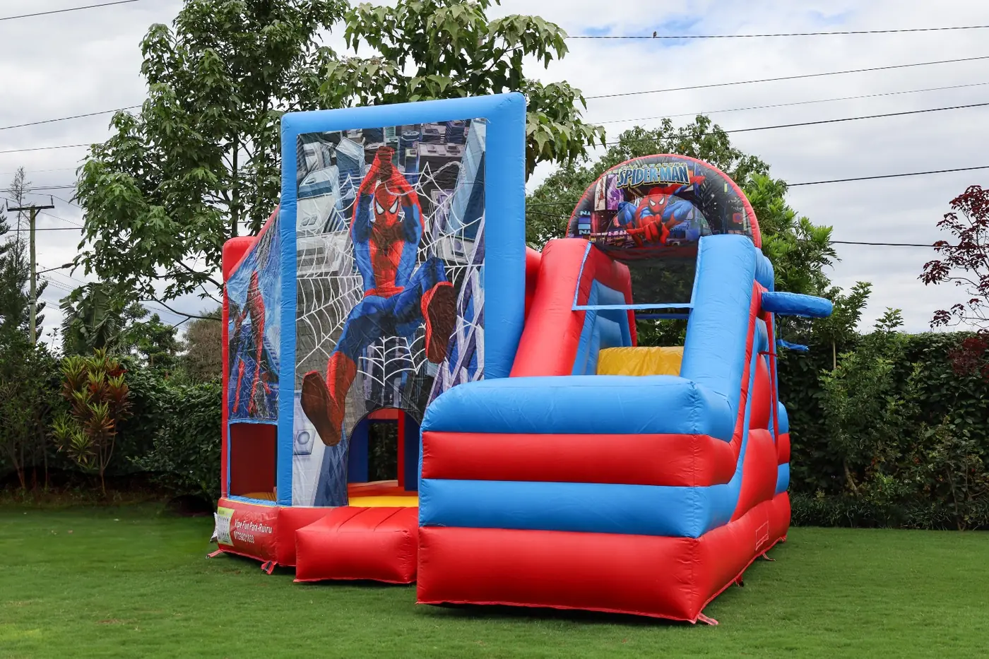 A wide view of a red and blue superhero-themed bouncing castle and slide on the lawn